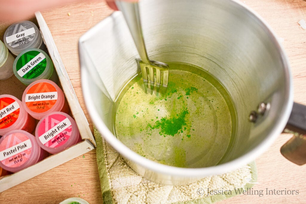 fork stirring green wax dye chips into hot wax in a metal pitcher to make wax melts