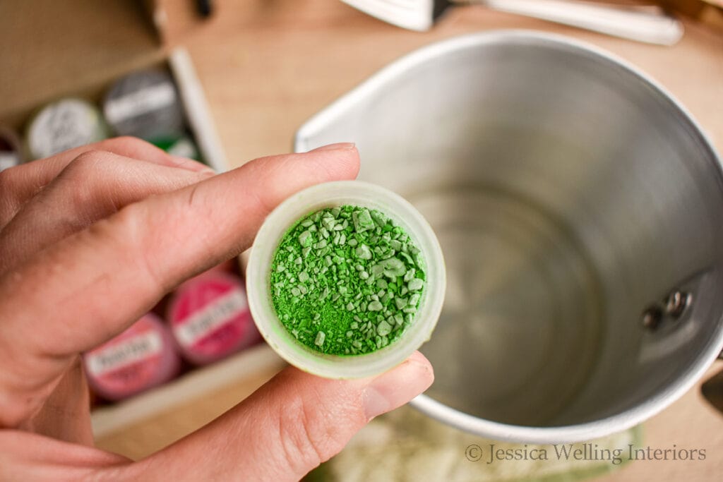 close-up of a container of green candle wax dye with a metal pitcher of melted wax in the background