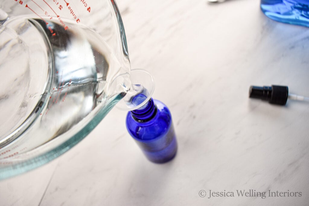 distilled water being poured into a small blue glass spray bottle to make DIY poo-pourri