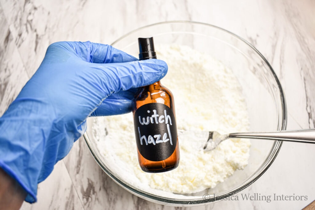 a gloved hand holding a small spray bottle of witch hazel over a bowl of bath bomb mix