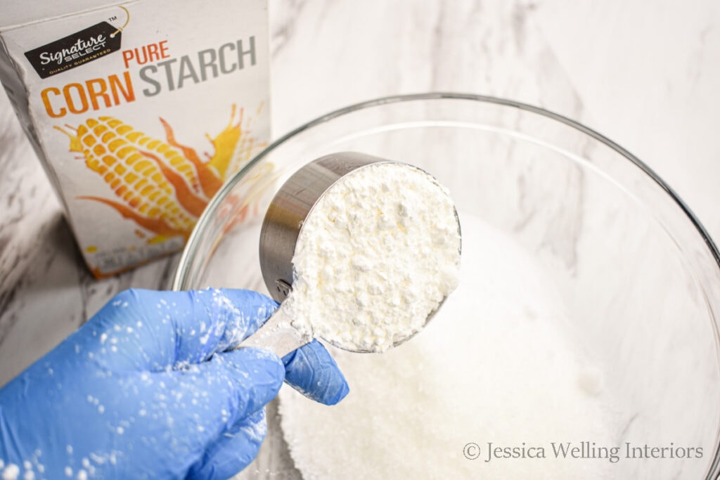 gloved hand pouring corn starch into a mixing bowl to make DIY bubble bath bombs