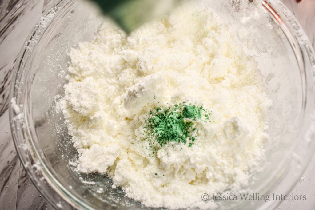 bowl of bath bomb mixture with green mica powder being sprinkled into it to add color