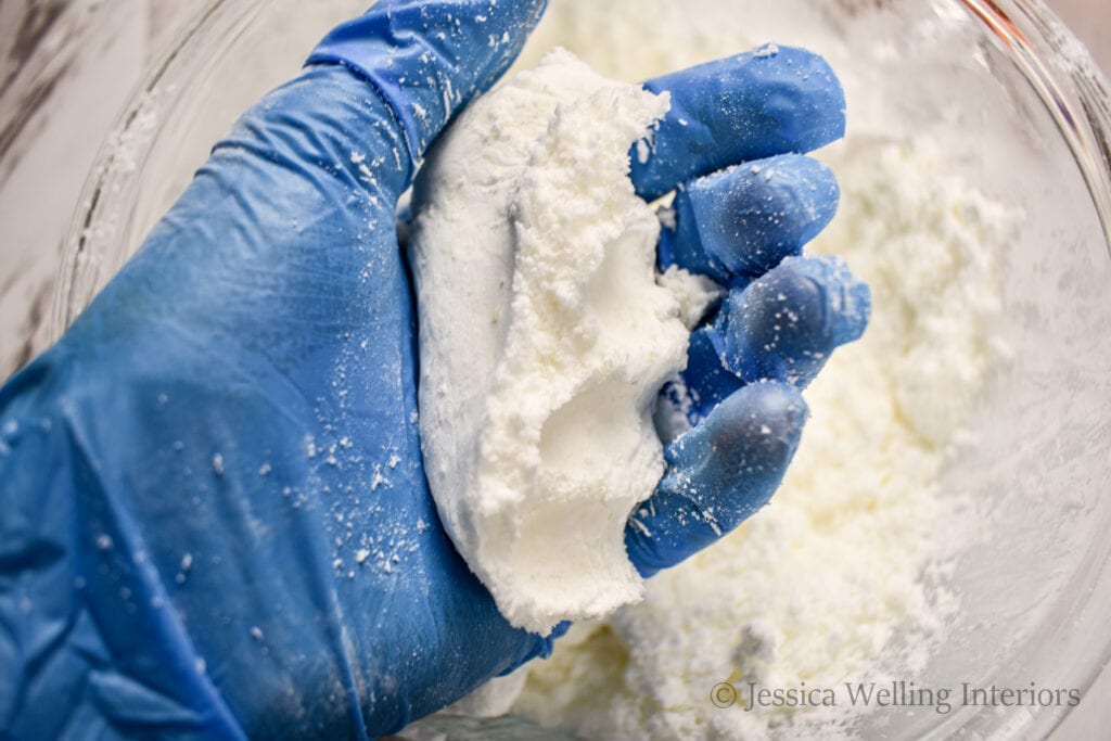 a gloved hand holding a pressed clump of bath bomb mixture to show its consistency