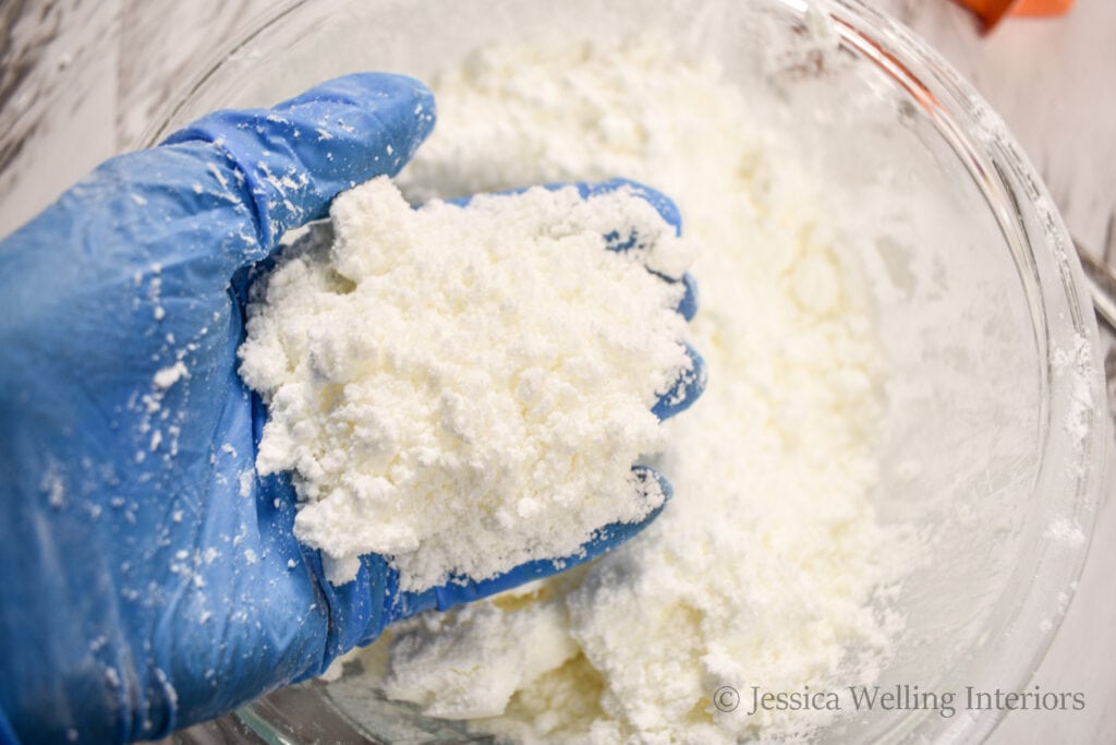 a gloved hand holding some bath bomb mixture over a mixing bowl to show its texture