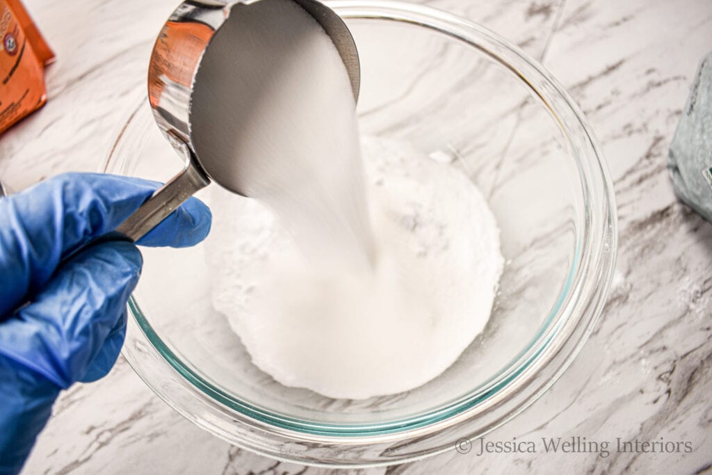 gloved hand pouring citric acid into a mixing bowl to combine the dry ingredients in the bath bomb recipe