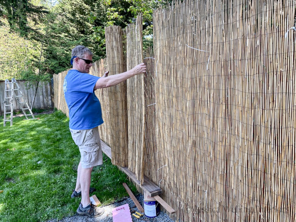 man holding a roll of bamboo reed fencing up against a fence