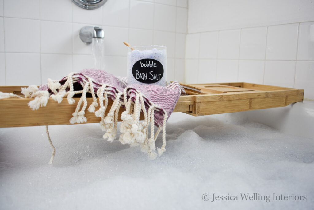 glass jar of homemade bubble bath salt sitting on a bath tray over a tub full of bubbles