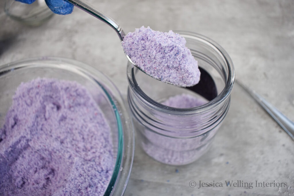 purple bath salt being spooned into a glass jar for storage