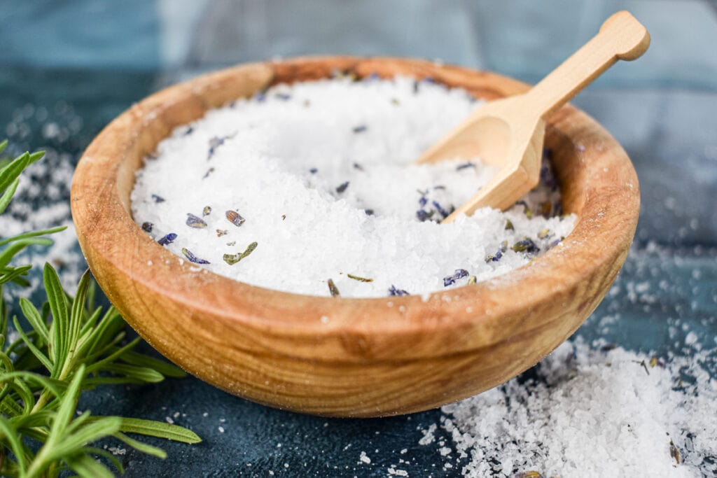 wood bowl of lavender scented bath salts on a tile countertop with a wood scoop and lavender sprigs