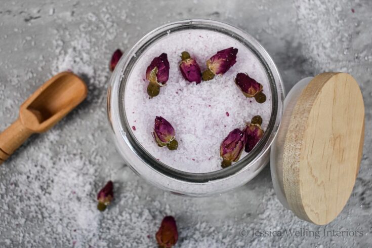 rose scented bath salts in a glass jar with rose buds and a wood scoop